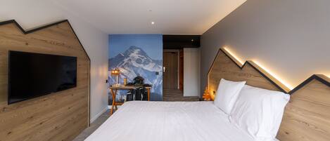 A modern hotel room with a large bed, a flat-screen TV, a desk with a chair, and a mountain mural on the wall.