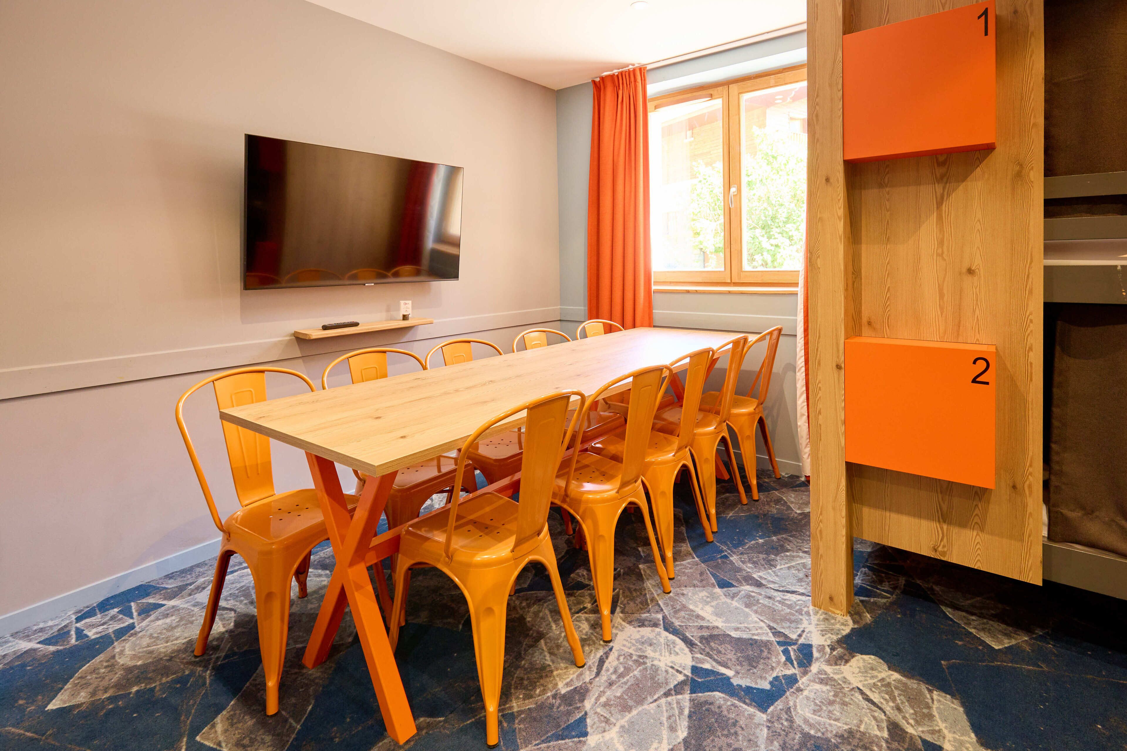 A conference room with a long table, orange chairs, a flat-screen TV, and a window with orange curtains.