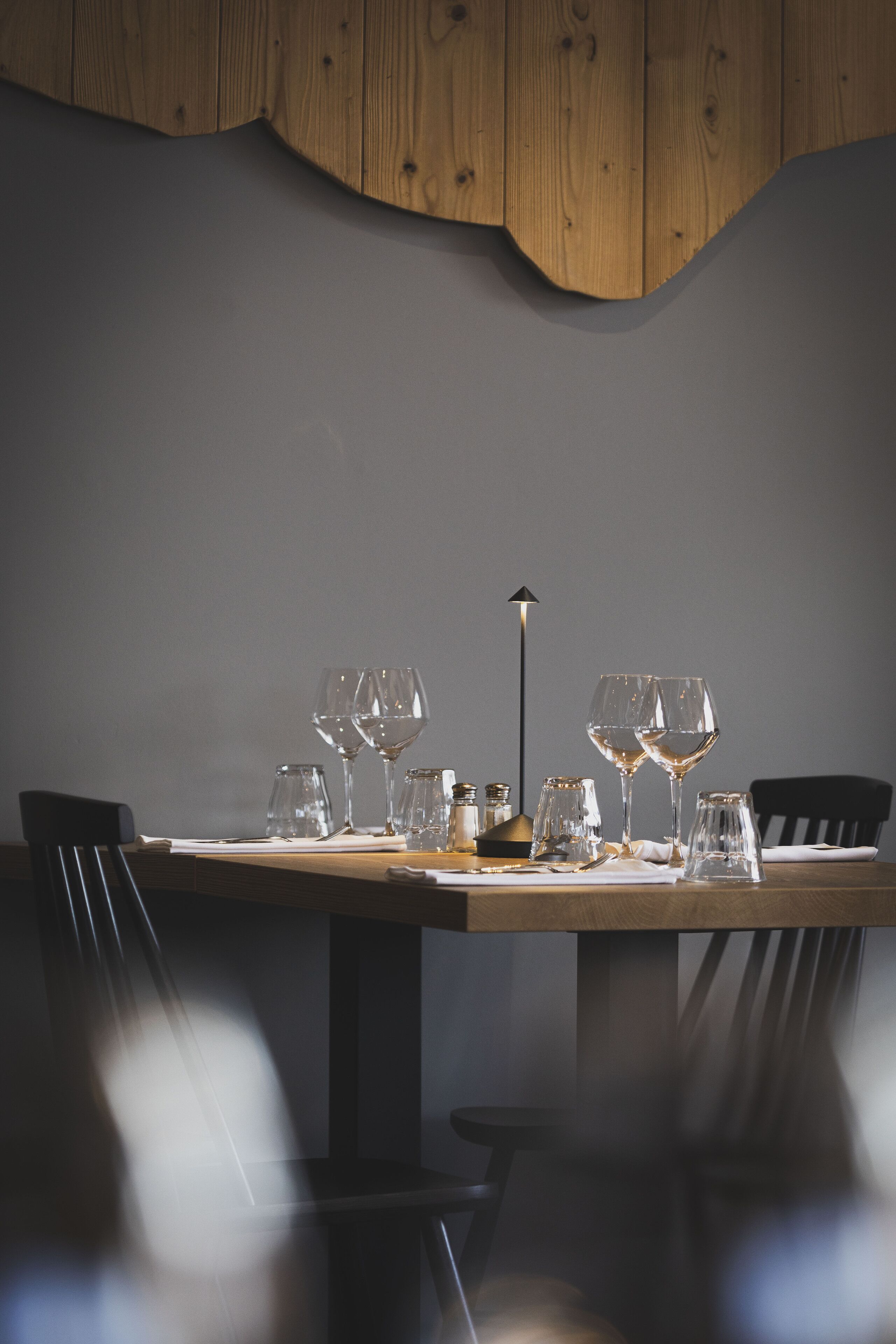 A dining table with empty wine glasses and a candle, set against a wooden wall and dark floor.