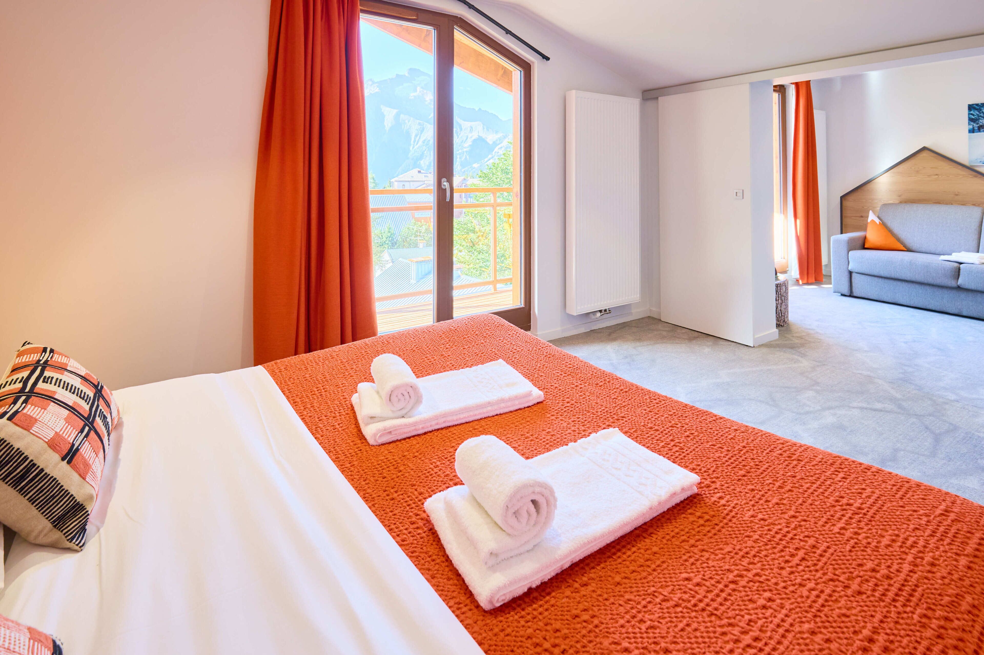 Een hotelkamer met een bed, twee opgerolde handdoeken, een bank en uitzicht op bergen door een groot raam.