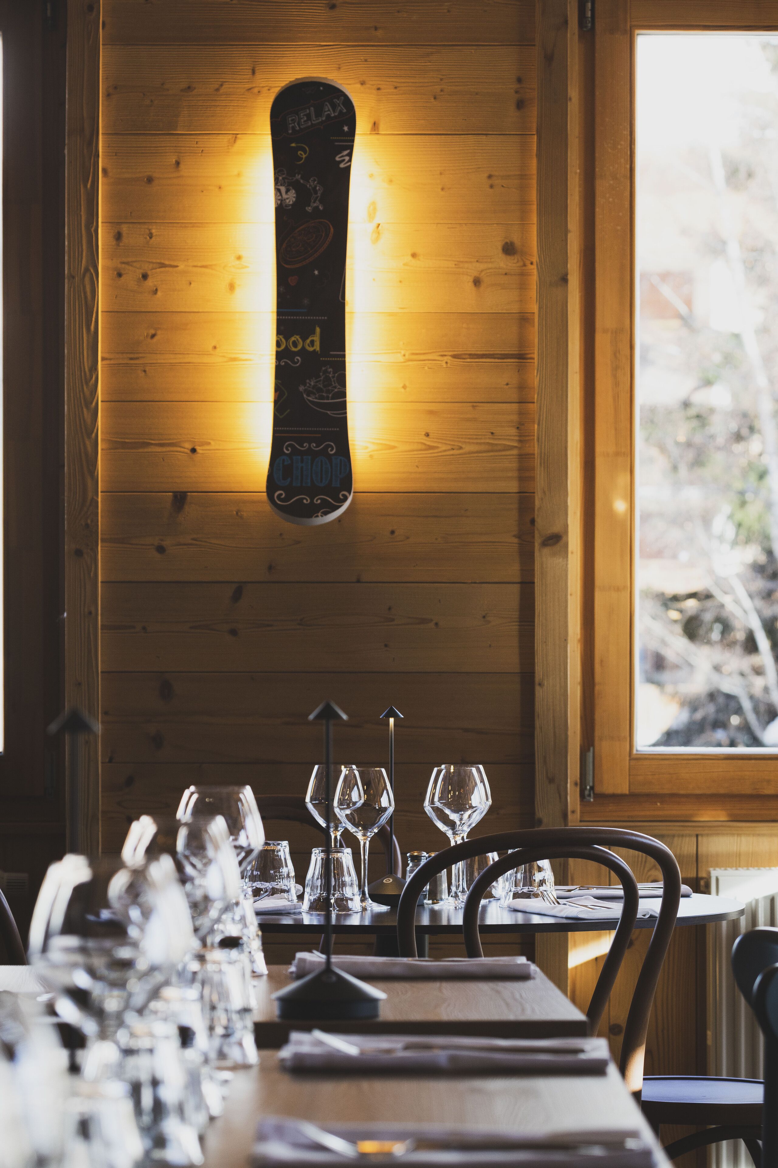 A wooden interior with a snowboard decoration, a dining table set with wine glasses, and a window with a view of trees.