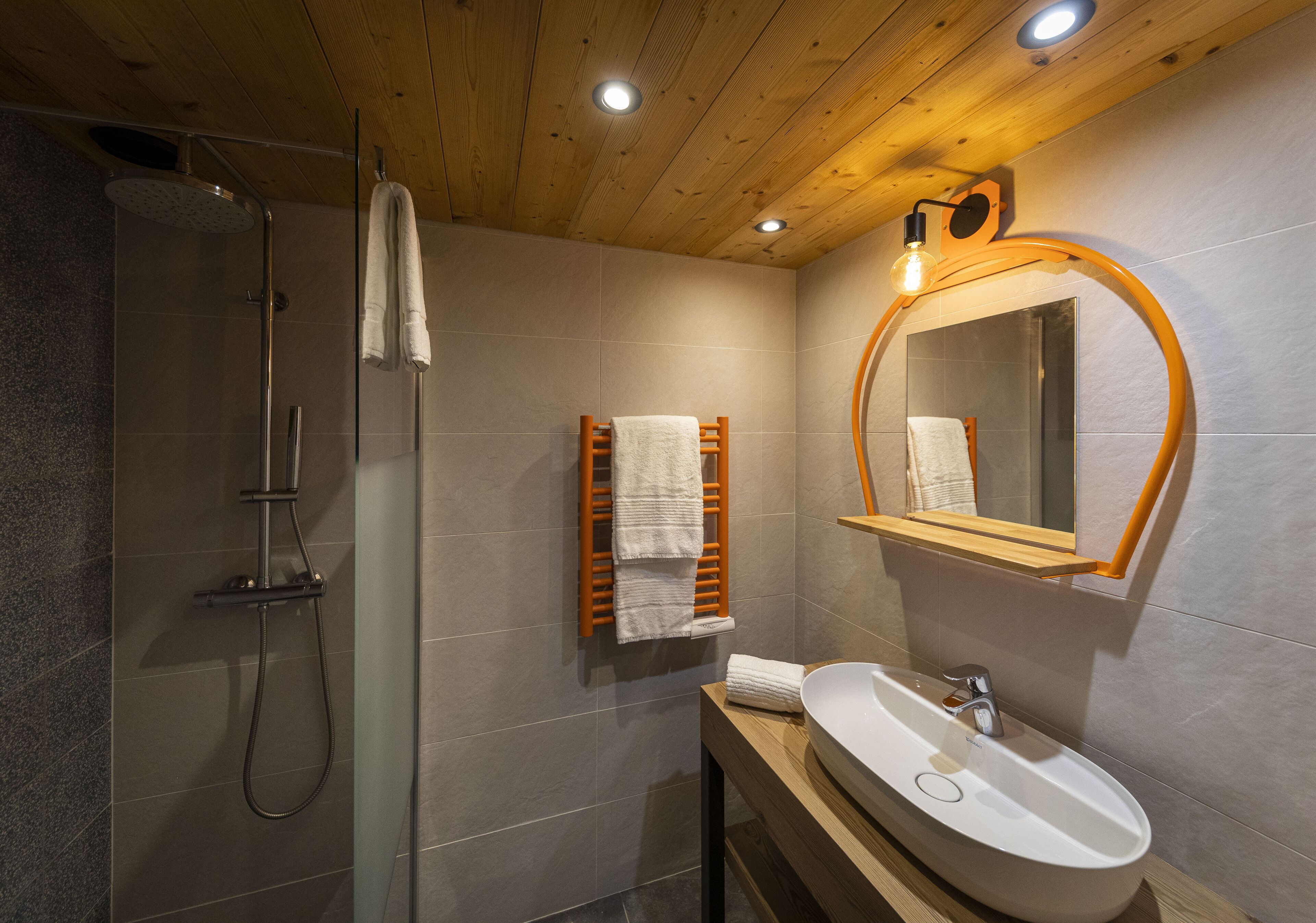Une salle de bain moderne avec une douche à parois de verre, un meuble-lavabo en bois avec un lavabo blanc et un miroir avec un cadre orange.