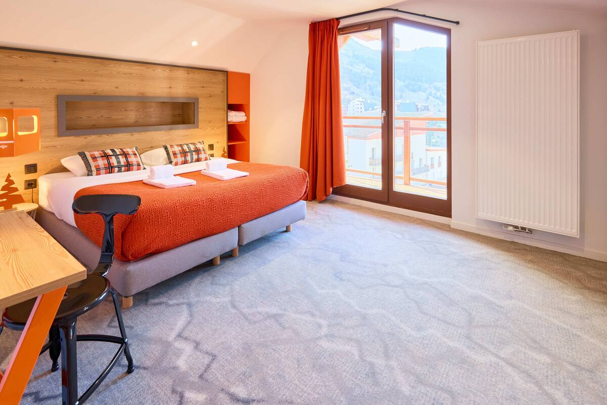 Une chambre d’hôtel avec un lit, un bureau, une chaise, une fenêtre donnant sur une montagne et un radiateur.