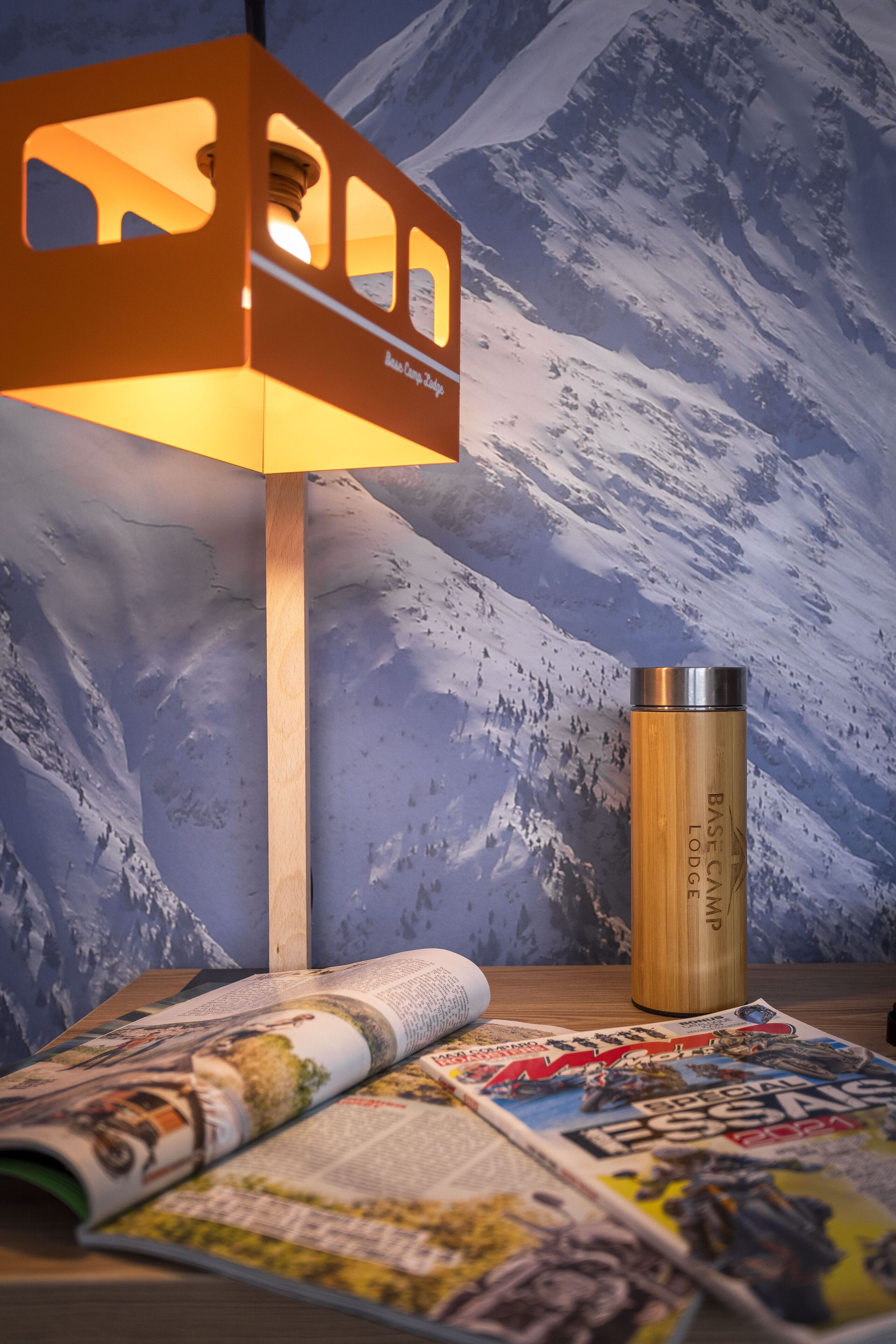 A room with a mountain-themed wall, a wooden table with a magazine, a lamp, and a stainless steel thermos.