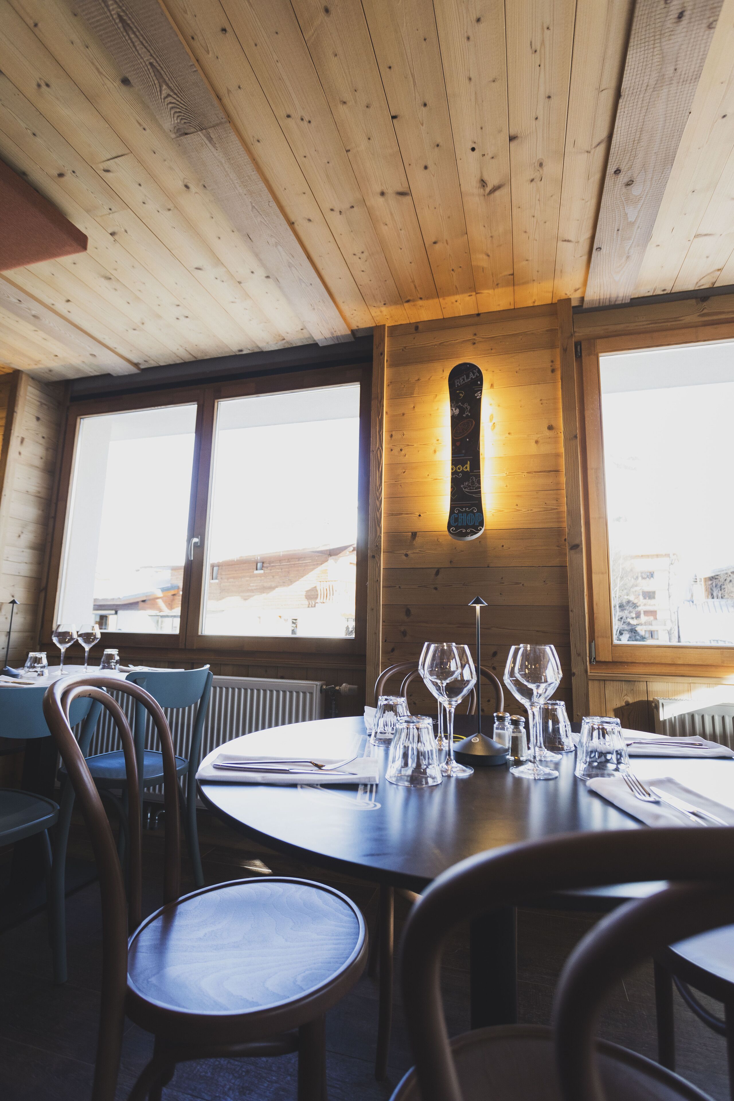 A wooden interior with a round dining table set for two, large windows, and a wall-mounted light fixture.