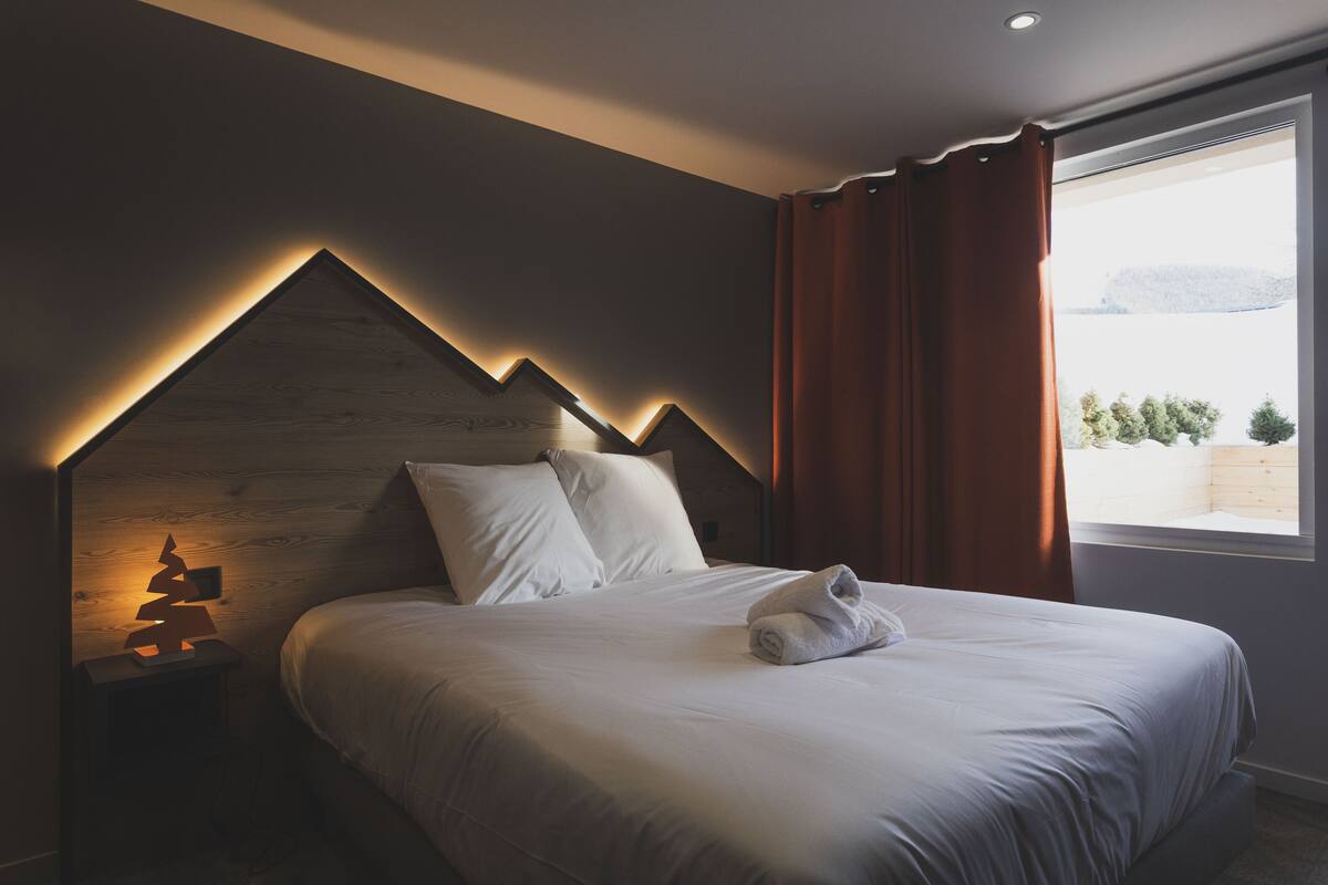 Une chambre à coucher avec une tête de lit en bois, une table de chevet avec une lampe, un lit bien fait et une fenêtre avec des rideaux.