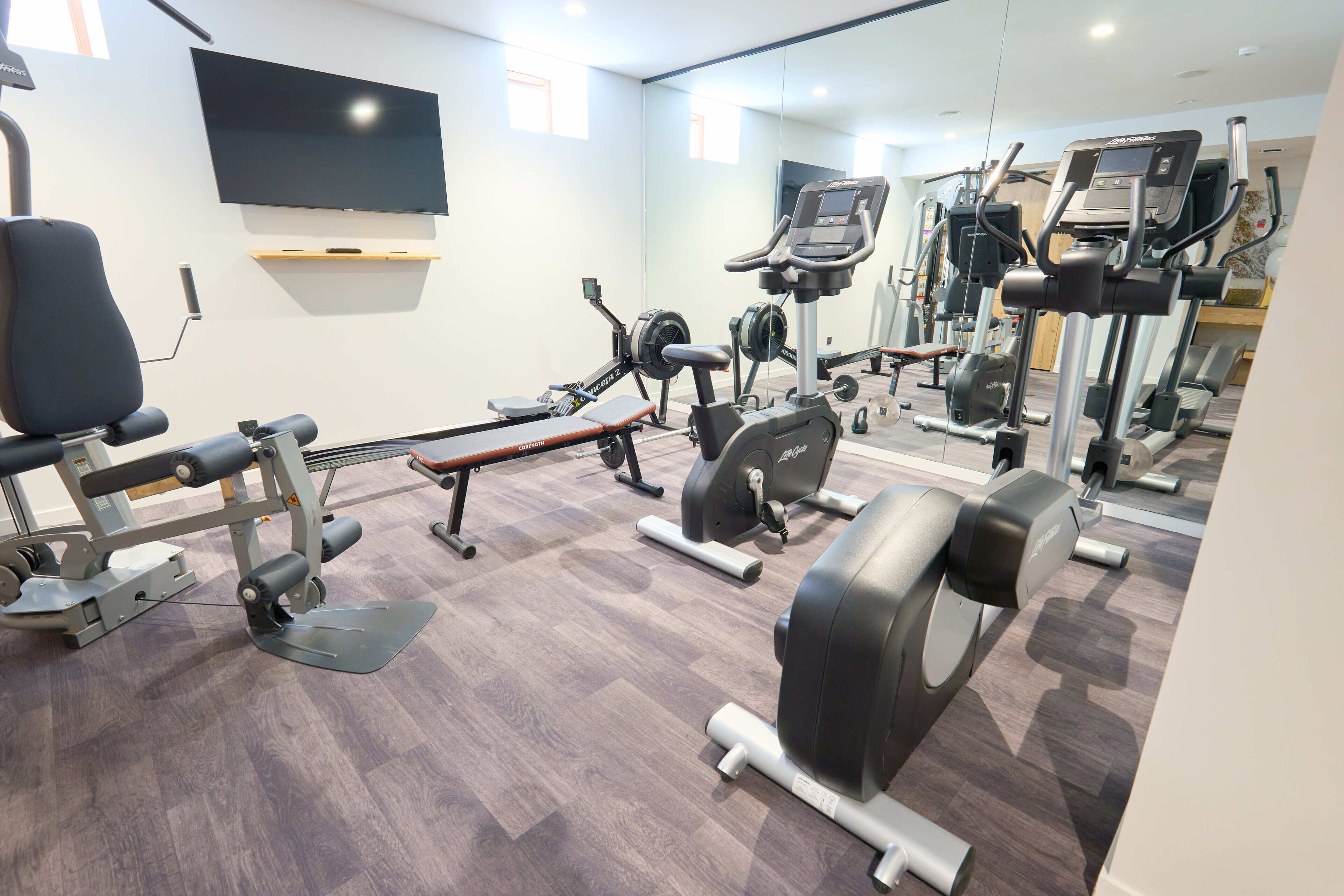 A gym with various exercise equipment, a television on the wall, and a mirror reflecting the equipment.