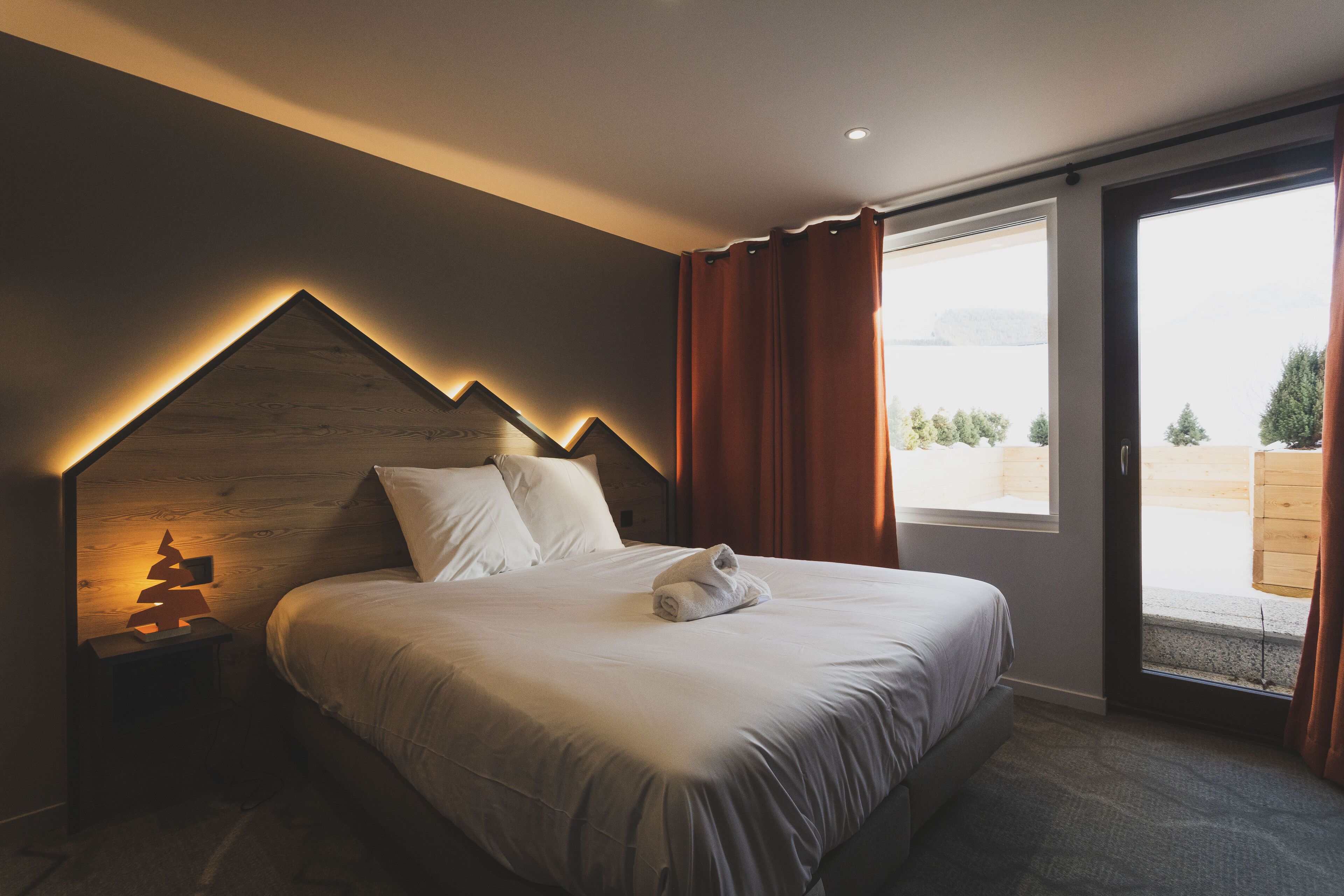 Ein Schlafzimmer mit einem Bett, einem Nachttisch, einem Fenster mit Vorhängen und Blick auf Bäume und Berge.