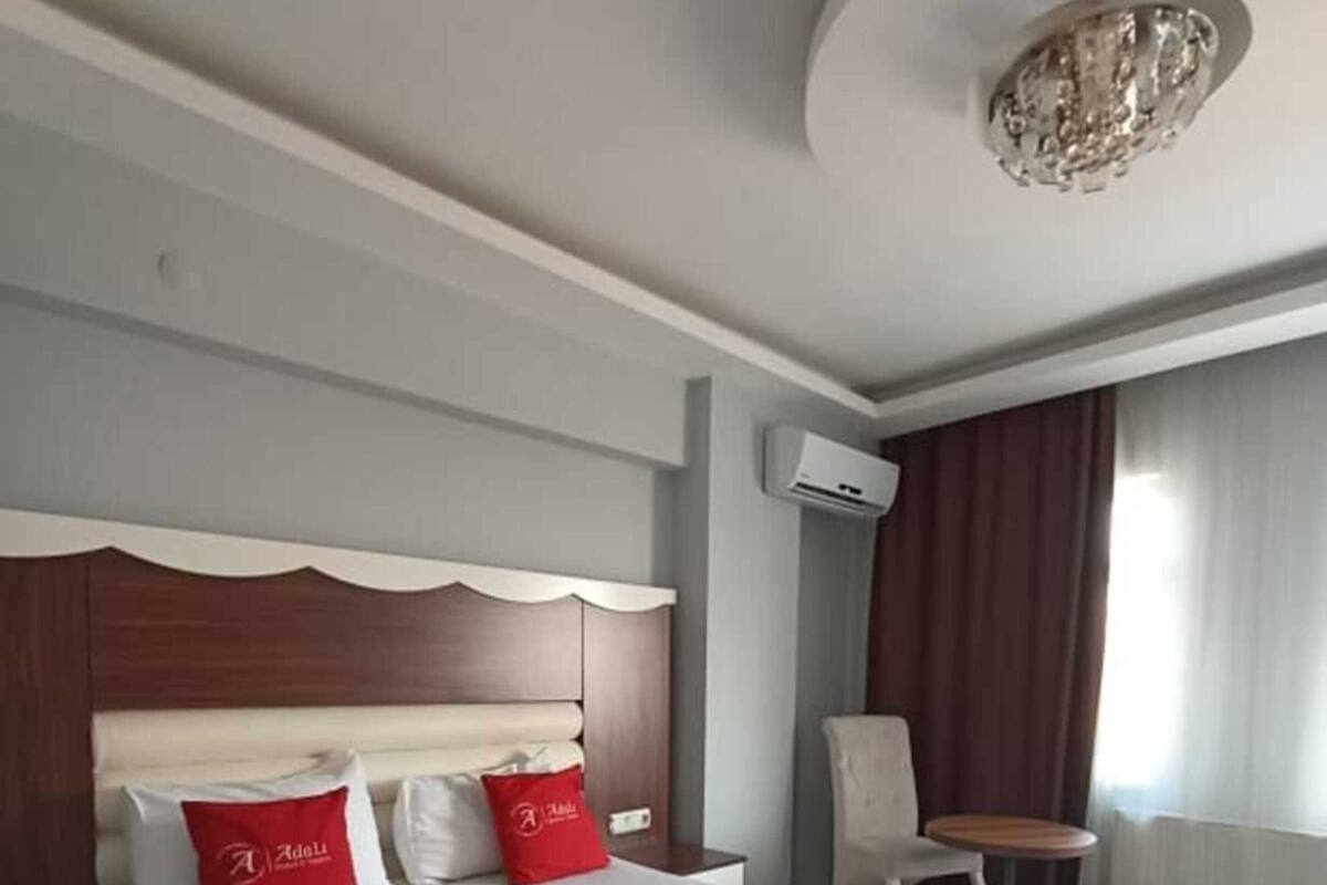 A hotel room with a large bed, a wooden headboard, a red pillow, a brown throw blanket, a small table, and an air conditioning unit.