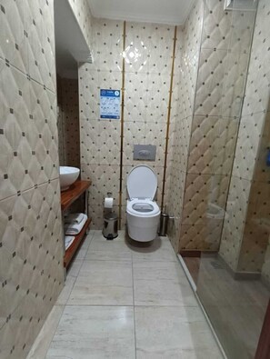 A small, tiled bathroom with a toilet, a shelf with towels, and a trash can.