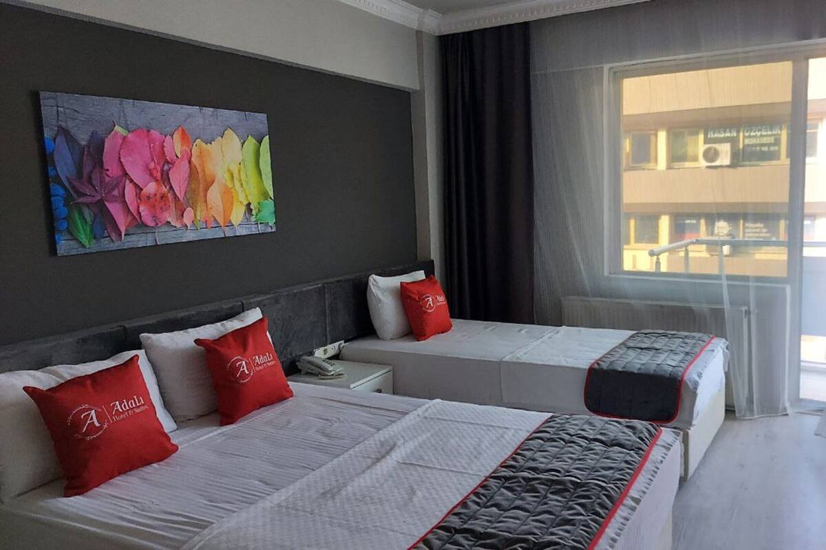 A hotel room with two beds, each with a red pillow and a white bedspread. There is a large window with a view of a building outside.