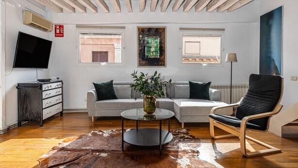 A modern living room with a sofa, a chair, a coffee table, and a TV. There are windows, a radiator, and a fire extinguisher visible. - Waou Puerta del Sol II - Alquiler Corta Duración (Madrid)