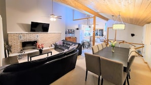 A modern living room with a fireplace, a black leather sofa, a dining table with chairs, and a kitchen in the background.