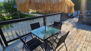 A wooden deck with a glass table and chairs, a thatched umbrella, and a white picket fence.