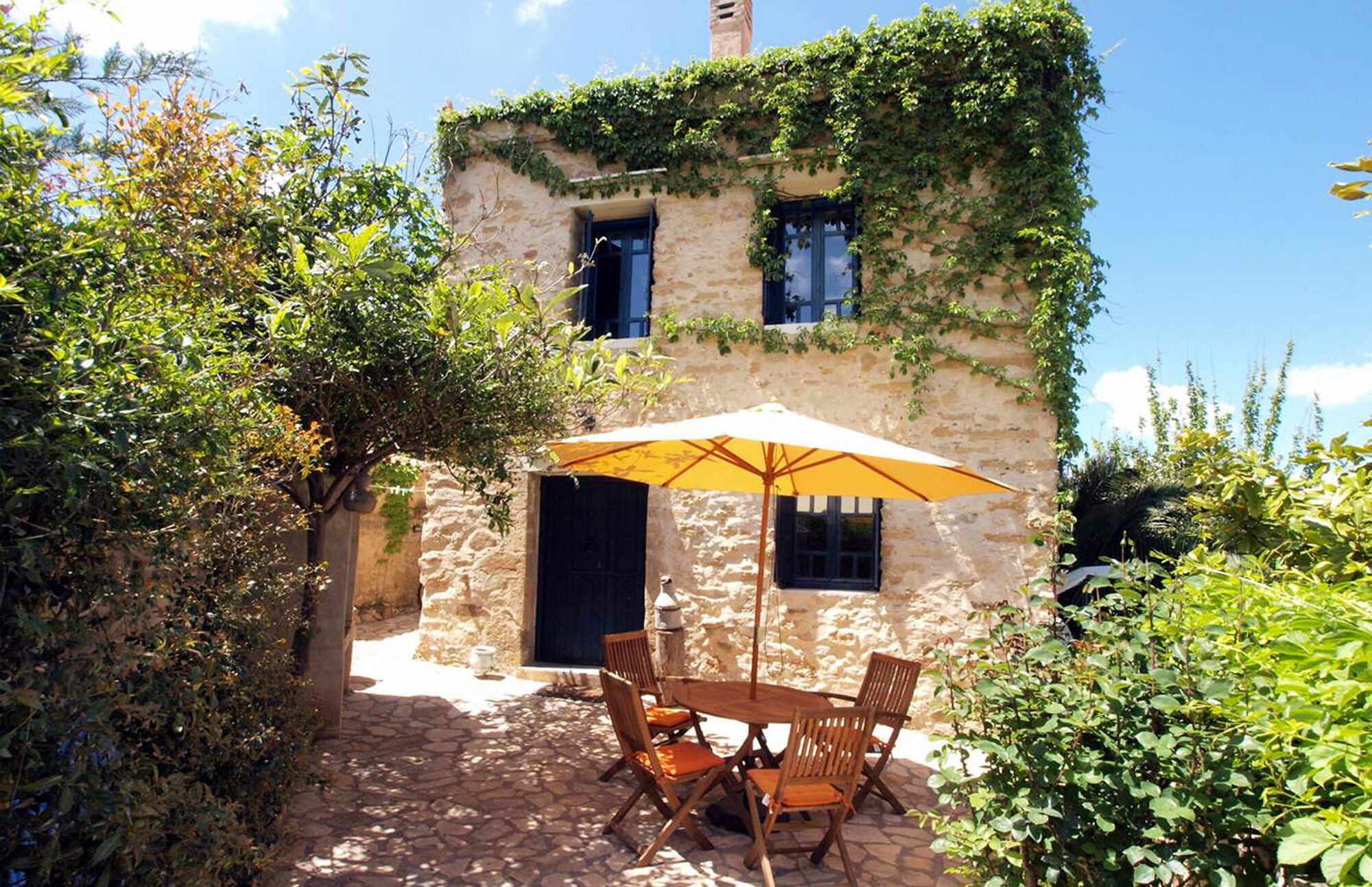 A stone building with ivy-covered walls, a wooden table with chairs, and a yellow umbrella.