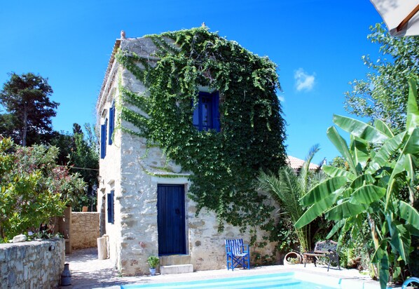 A stone building with a blue door and window, partially covered in ivy, with a pool and outdoor seating area.