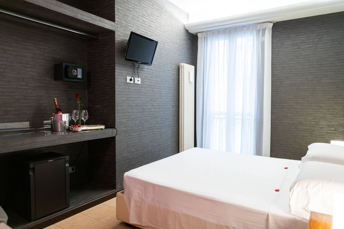 A hotel room with a bed, a TV mounted on the wall, a small fridge, a bedside table with a bottle and glasses, and a window with curtains.
