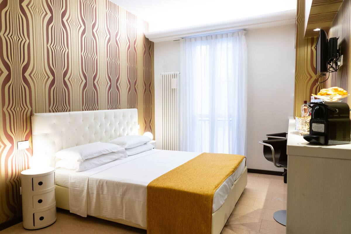 A hotel room with a large bed, a bedside table, a desk with a chair, a radiator, and a television mounted on the wall.