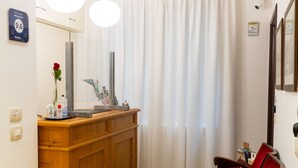 A compact living space with a wooden cabinet, a small table with a vase, a black patterned rug, and a window with white curtains.