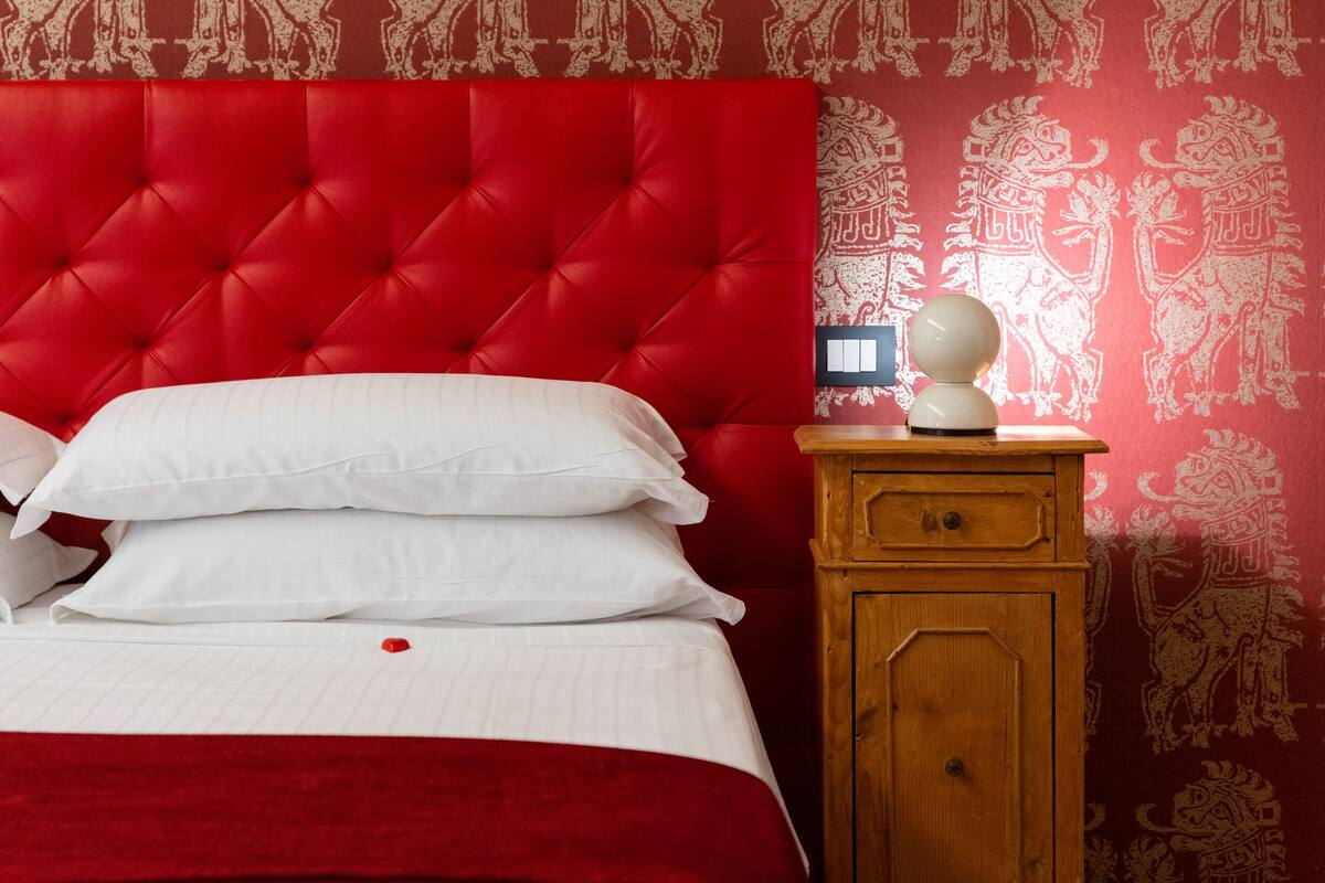 A room with a red upholstered headboard, a wooden nightstand with a lamp, and patterned wallpaper.