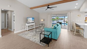 A spacious living room with a teal sofa, a glass coffee table, and a ceiling fan. A TV is mounted on the wall, and a window offers a view of a pool and outdoor area. - Look at this View with direct Gulf Access (Cape Coral)