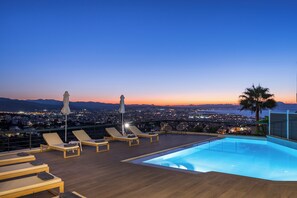 A rooftop pool with lounge chairs and umbrellas, overlooking a cityscape at dusk.