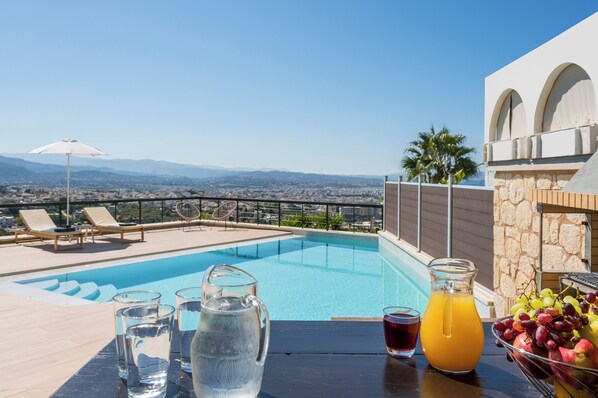 A poolside table with drinks and a fruit bowl, overlooking a city and mountains.