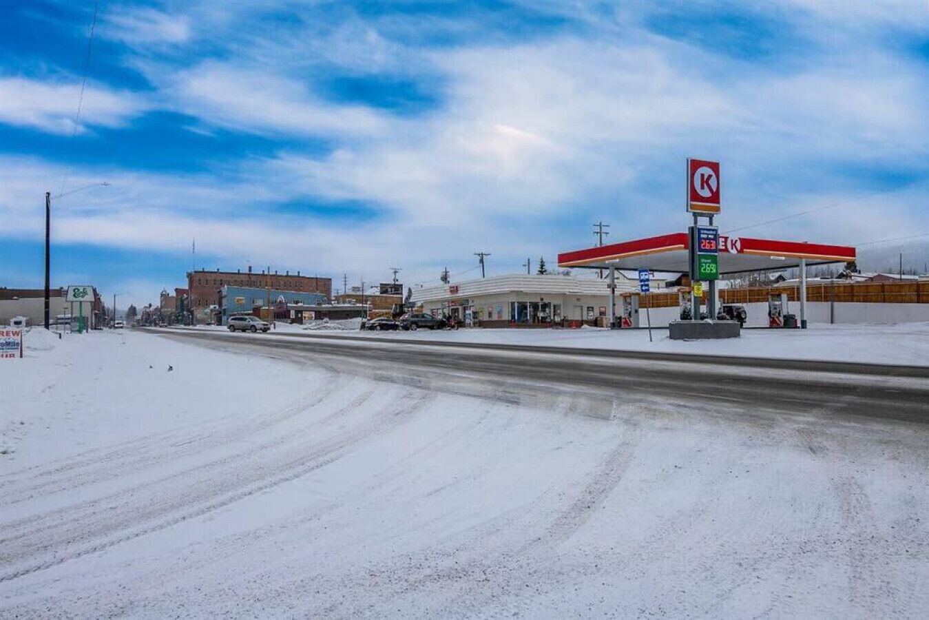 Tankstelle mit roter Markise und dem 'K'-Logo, umgeben von Gebäuden und einer verschneiten Landschaft.