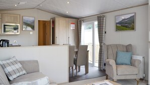 A cozy living room with a sofa, a chair, a coffee table, and a kitchen area with a kettle. - Tranquil Lodge - UK35277 (Brigham, near Cockermouth)