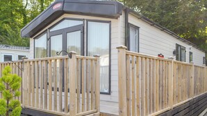 A mobile home with a wooden deck, a small porch, and a window with a view of the surrounding area. - Tranquil Lodge - UK35277 (Brigham, near Cockermouth)