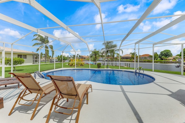 A covered outdoor swimming pool with two wicker lounge chairs and a concrete deck.