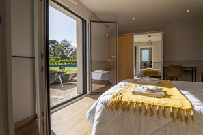 Een slaapkamer met een grote schuifpui naar een tuin, een bed met wit beddengoed en een gele plaid, en een spiegel die de kamer reflecteert.