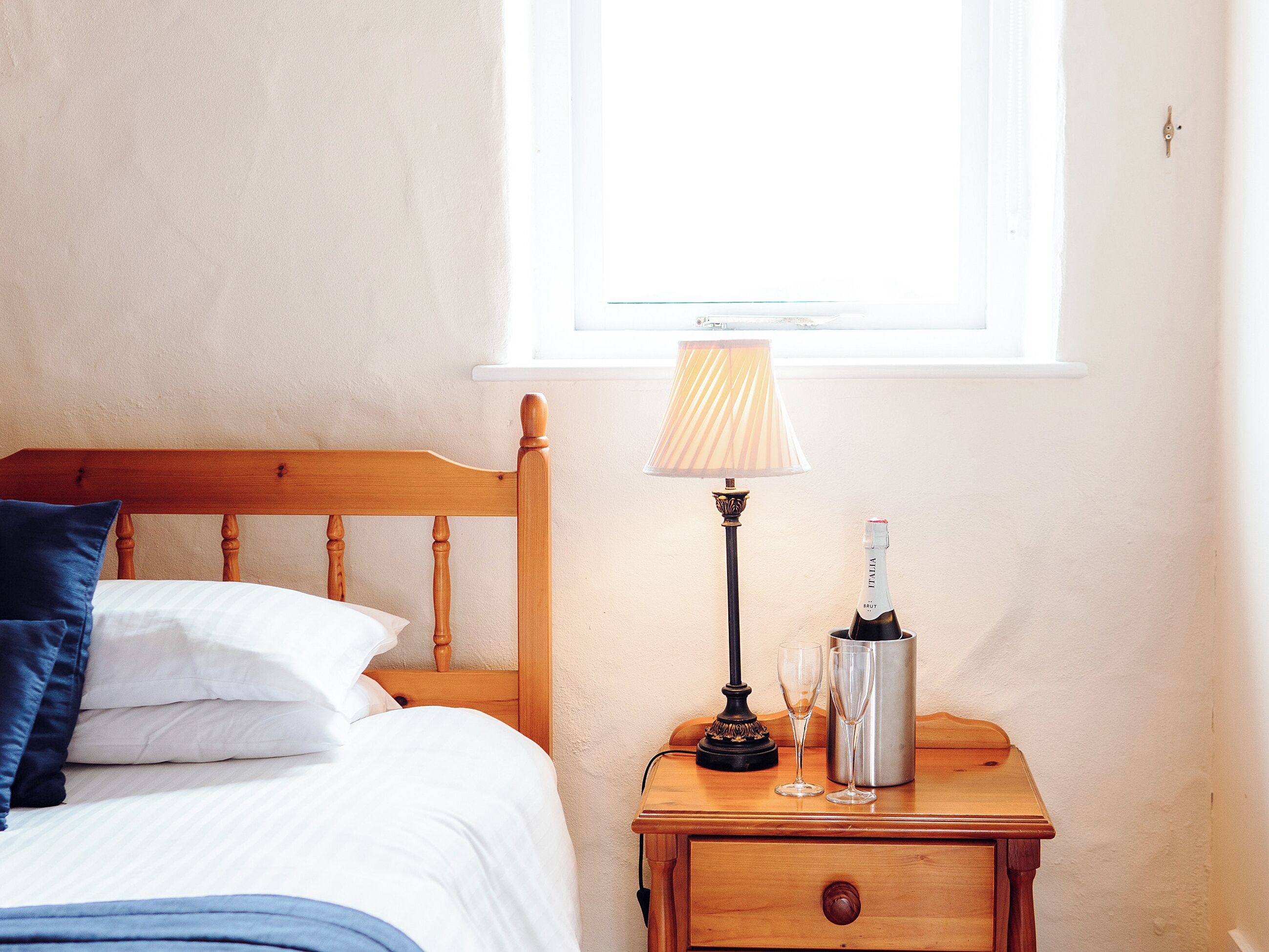 A neatly made bed with pillows, a bedside lamp, a bottle of champagne, and a glass on a wooden nightstand.