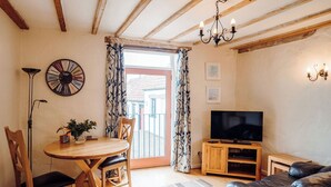 A living room with a wooden dining table, two chairs, a TV stand, a leather sofa, a chandelier, and a large window with curtains. - The Parlour - UK6717 (Lydstep, near Tenby)