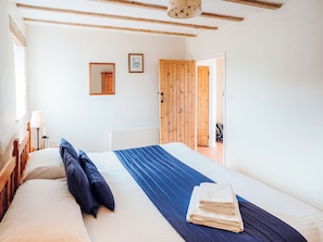 A bedroom with a bed, pillows, a blue blanket, a folded towel, a nightstand with a lamp, a framed picture, and a wooden door.