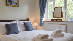 Un lit bien fait, avec du linge de lit blanc et des coussins à motifs bleus, une lampe de chevet, une fenêtre donnant sur des arbres et une chaise en bois.