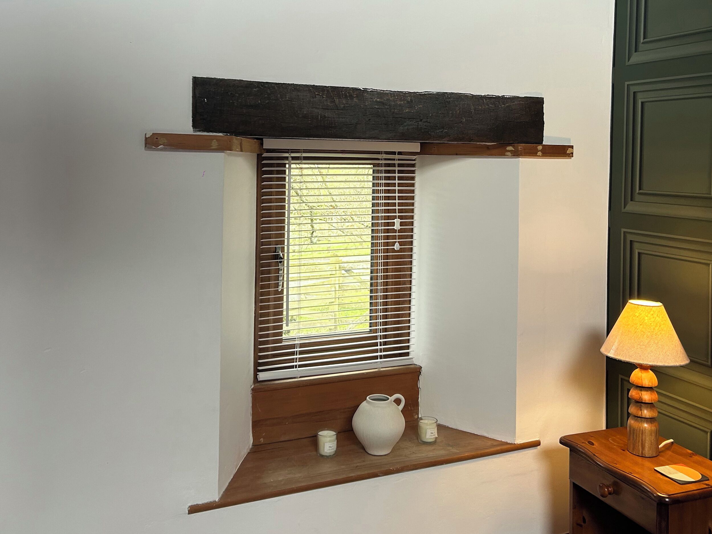 Una habitación con ventana y persianas, un estante pequeño con un florero y velas, y una lámpara sobre una mesa de madera.