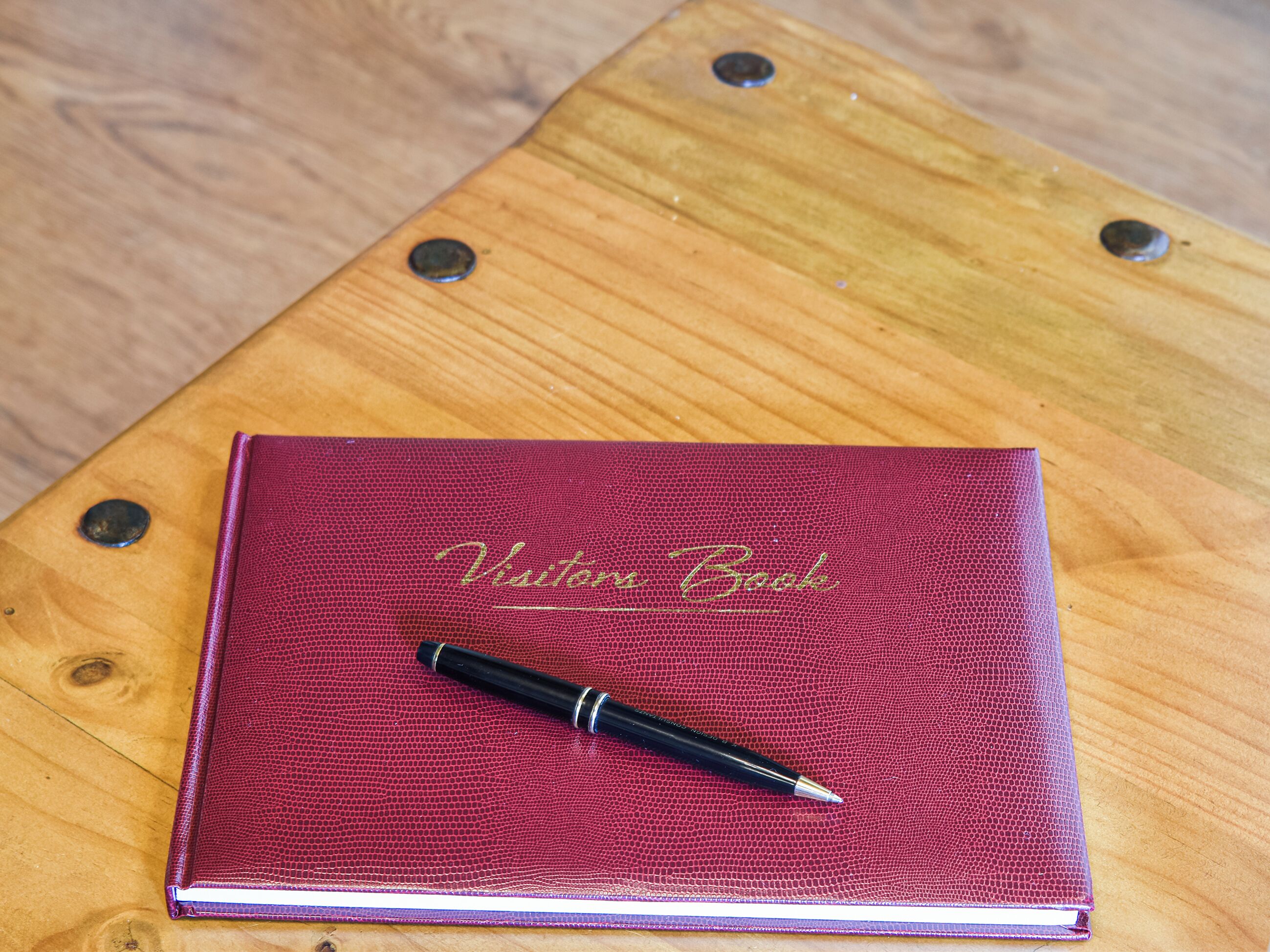 A red leather-bound book titled "Visitor Book" with a pen on top, placed on a wooden surface.