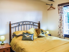 Een bed met een metalen hoofdbord, een nachtkastje met een lamp en een raam met bloemen gordijnen.