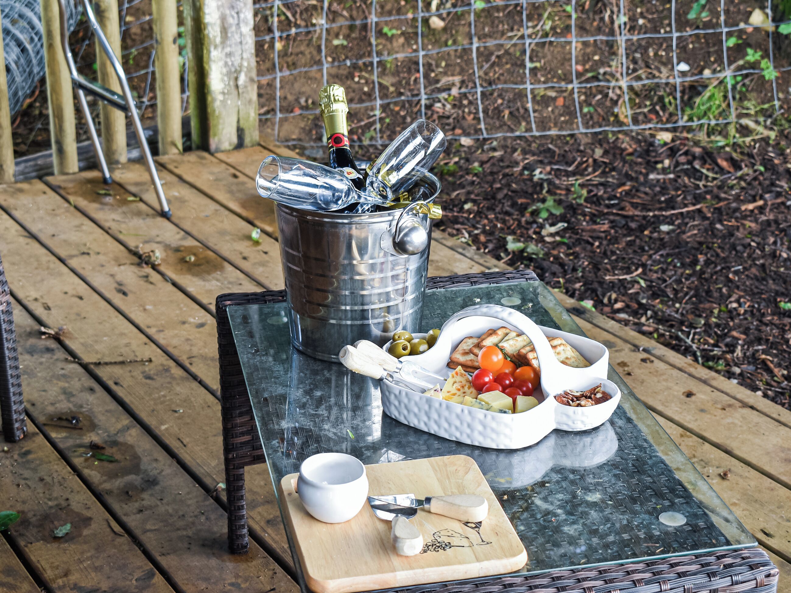 Een picknickopstelling op een houten terras, met een metalen emmer, een witte schaal, een snijplank en een beker.