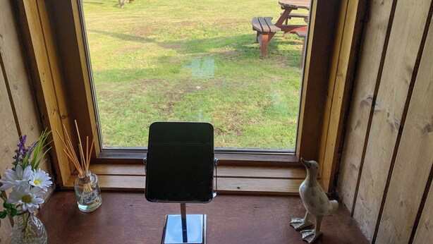 A room with a view of a grassy field and the ocean through a large window. There is a wooden bench outside the window, a small table with a chair inside, and a decorative bird statue on the windowsill. - Mains of Auchmedden Cottage (Near Pennan)