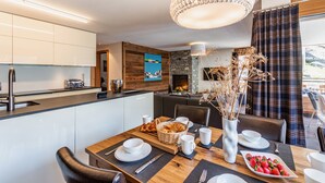 A modern dining area with a wooden table set for breakfast, a kitchen in the background, and a large chandelier. - Luxurious Chalet Petit - matterhorn views, central location, outdoor hot tub (Zermatt)