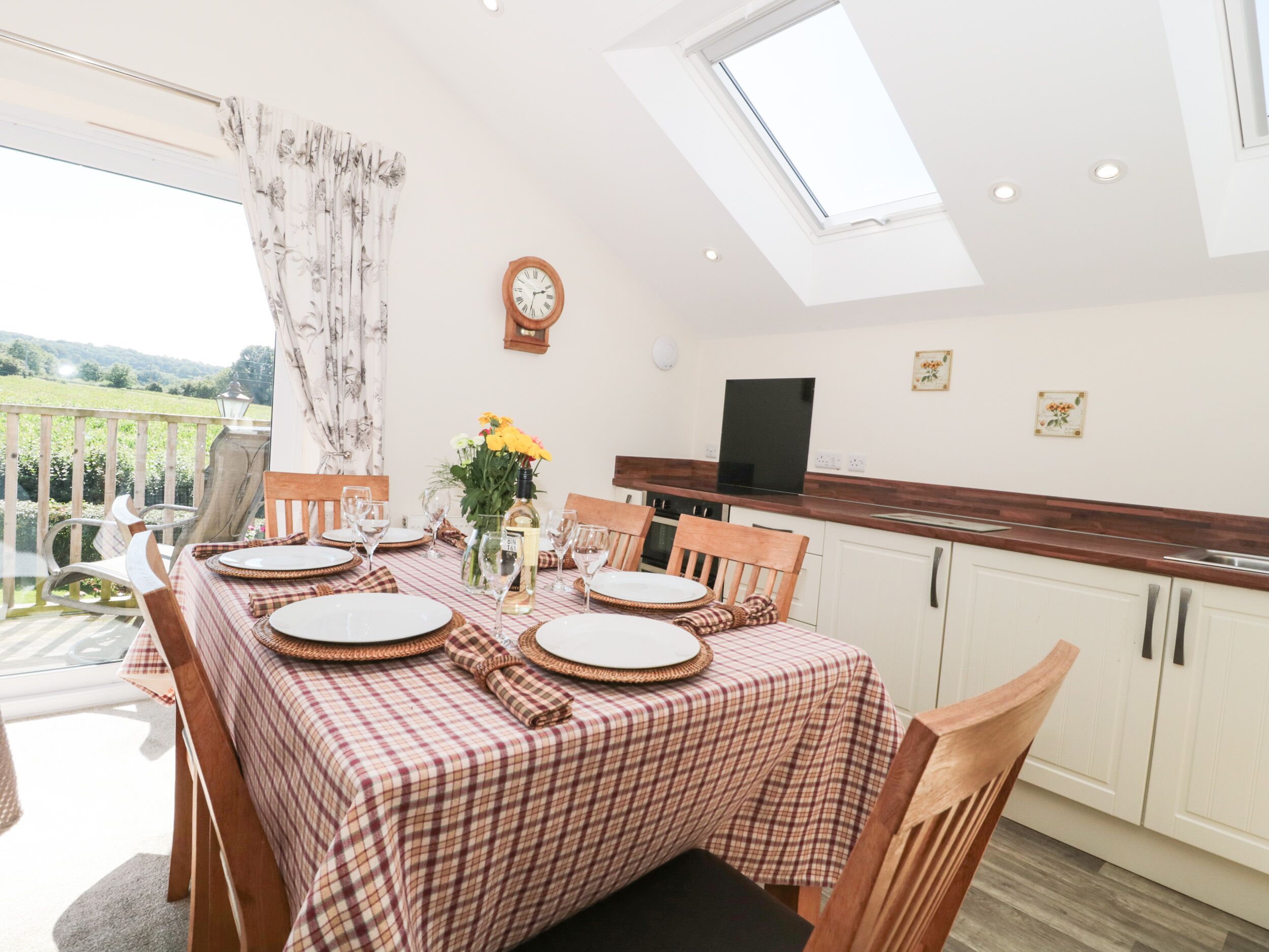 A dining area with a checkered tablecloth, wooden chairs, a clock, and a TV on the wall.