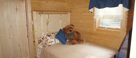 Ein Schlafzimmer in einer Holzhütte mit einem Einzelbett, einem Fenster mit Vorhängen und einem Teddybären auf dem Bett.