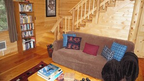 Interior de fusta amb una escala, un sofà amb coixins estampats, una biblioteca i una taula de centre amb llibres.