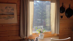 A wooden cabin interior with a window, curtains, a white chair, a table, a vase with flowers, a mug, and a framed picture. - Beautiful Mountain Cabin near Tetons Yellowstone (Dubois)