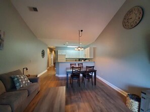 Amplia sala de estar con sofá y comedor con sillas y una mesa. Se ve una cocina al fondo, con armarios y un reloj en la pared.