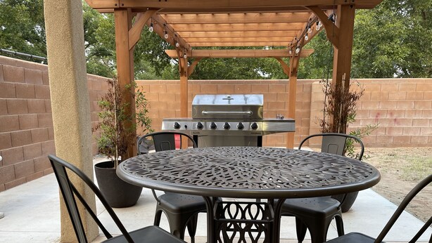 Une terrasse extérieure avec une table ronde en métal et quatre chaises, un barbecue et une pergola décorée de guirlandes lumineuses.