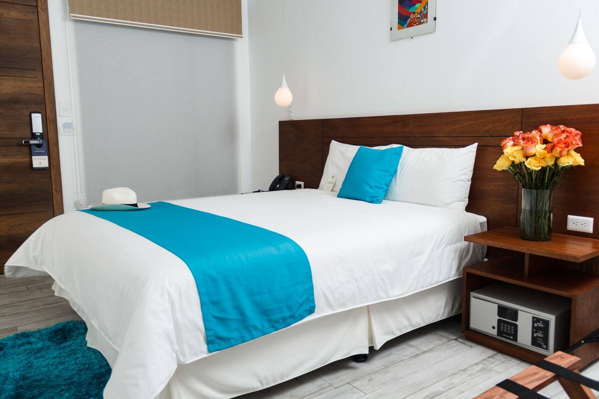 Une chambre d’hôtel moderne avec un lit bien fait, une table de chevet ornée d’un vase de fleurs et une petite armoire équipée d’une prise électrique.