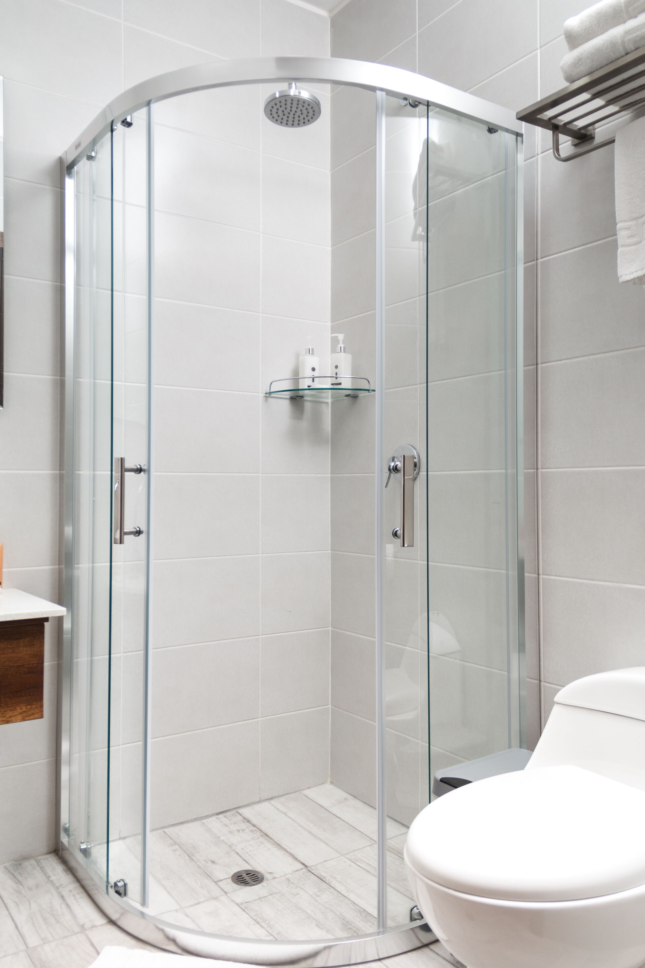 Un baño moderno con ducha acristalada, inodoro blanco y repisa de vidrio con artículos de aseo.